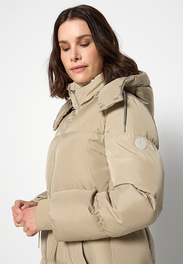 Femme portant une doudoune beige avec un col haut et une capuche, regardant vers le bas avec les mains légèrement jointes devant un fond uni.