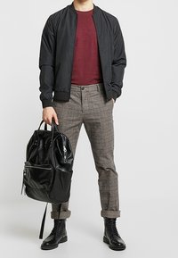 Osoba v černé bomber bundě, bordó košili, kostkovaných kalhotách, černých šněrovacích botách, držící černý kožený batoh.