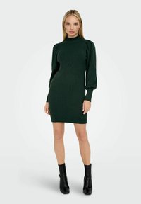 Robe en tricot vert foncé avec un col haut, texture à côtes, silhouette ajustée et manches bouffantes, associée à des bottines à talons noires.