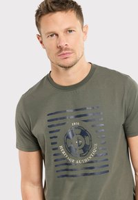 T-shirt en coton vert olive avec un col rond, des manches courtes et un design imprimé marine avec des rayures, un disque et du texte.