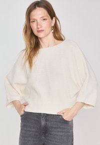 Pull en maille lâche de couleur crème avec de larges manches et un col rond, associé à un jean gris foncé. Le tissu présente des coutures texturées.