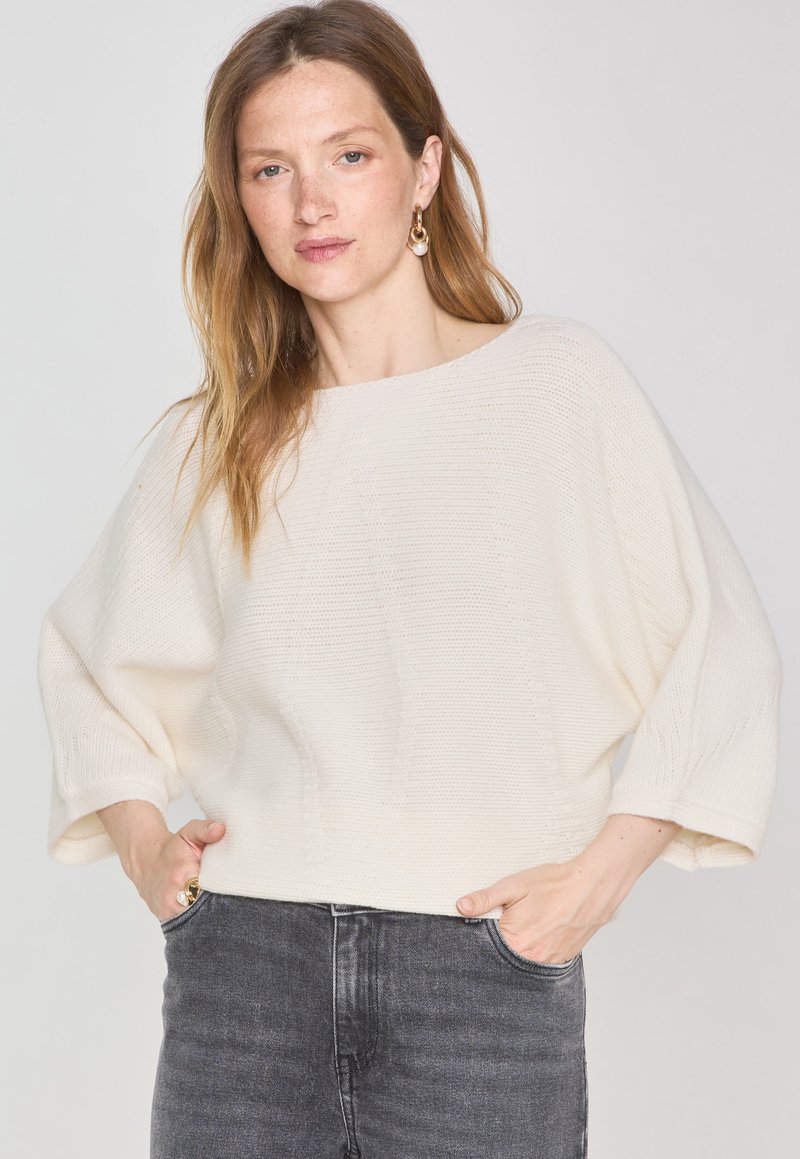 Pull en maille lâche de couleur crème avec de larges manches et un col rond, associé à un jean gris foncé. Le tissu présente des coutures texturées.