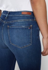 Vaqueros de mezclilla azul oscuro con un diseño ajustado, que cuentan con una etiqueta de cuero con "TOMMY HILFIGER" y un único bolsillo trasero con costura amarilla.