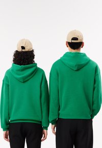 Sudaderas con capucha verdes, fabricadas en un material suave, que presentan un corte relajado y puños de canalé. Gorras beige llevadas hacia atrás completan el look.