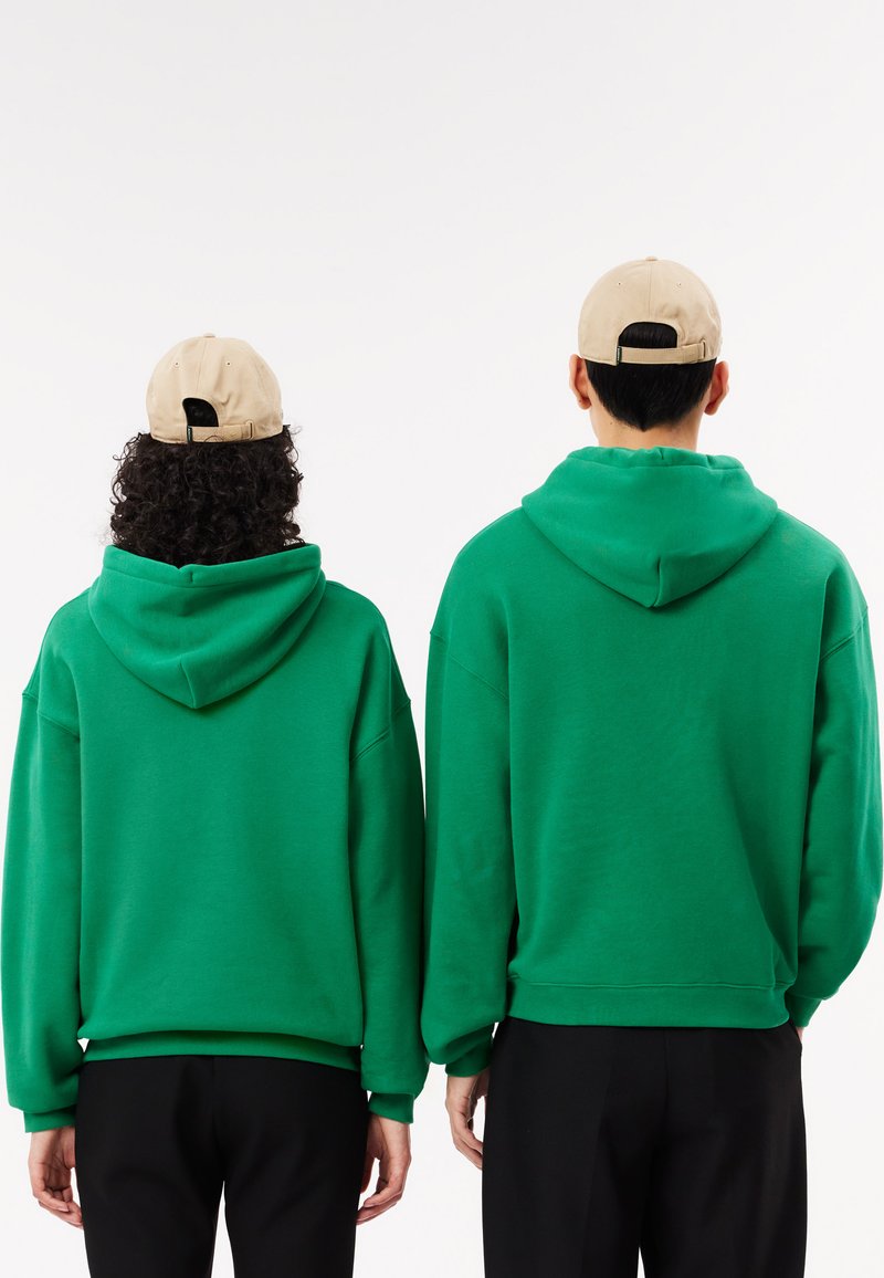 Sweatshirts à capuche verts en tissu doux, avec une coupe décontractée et des poignets côtelés. Des casquettes beiges portées à l'envers complètent le look.