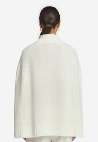 Vitt stickat poncho med ribbad nederkant och hög krage. Har en lös passform med texturerat mönster och sänkt axeldesign.