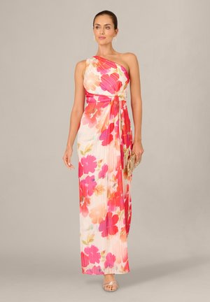 Kobieta ubrana w białą, asymetryczną maxi sukienkę z dużym różowo-pomarańczowym kwiatowym wzorem, trzymająca plecioną kopertówkę i nosząca srebrne sandały.