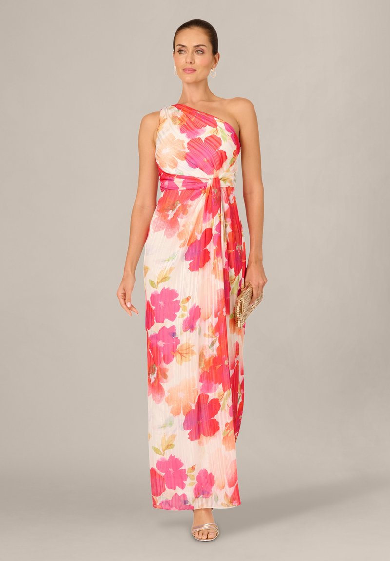 Vrouw draagt een witte maxi-jurk met één schouder en een grote roze en oranje bloemenprint, houdt een geweven clutch vast en draagt zilveren sandalen.