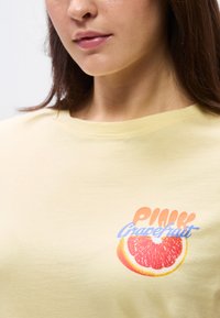 Γυναίκα που φοράει απαλό κίτρινο πουκάμισο με μια φέτα ροζ γκρέιπφρουτ και το κείμενο «Ροζ Γκρέιπφρουτ» στην περιοχή του στήθους.