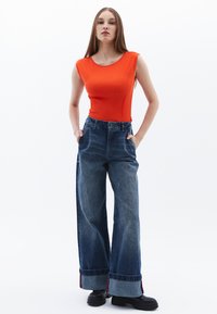 Top arancione senza maniche abbinato a jeans in denim blu a gamba larga con orlo risvoltato e dettagli scuri. Scarpe nere completano l'outfit.