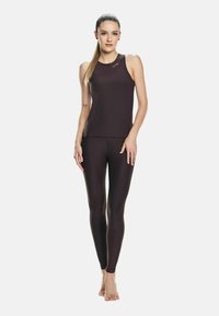 Donna in piedi a piedi nudi, indossa un top senza maniche nero e leggings coordinati, con un tatuaggio di farfalla sul suo avambraccio sinistro.