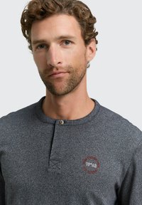 Dunkelgraues, strukturiertes Henley-Shirt mit einem runden Emblem, das "1962" zeigt. Der Kragen hat einen Knopf und ein dezentes Melange-Muster.
