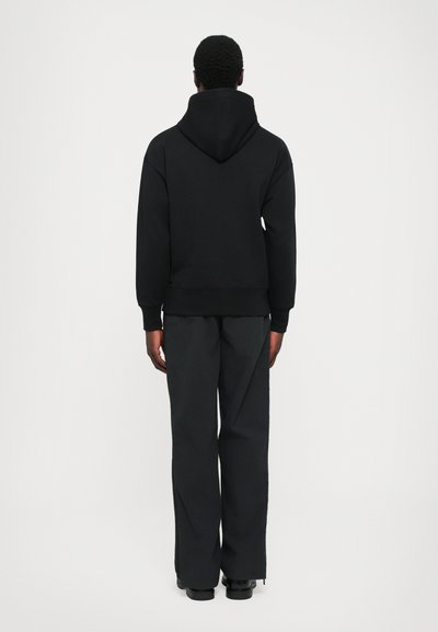 Uomo in piedi con la schiena rivolta, che indossa un cappuccio nero abbassato, pantaloni neri a gamba larga e scarpe nere, su uno sfondo bianco semplice.