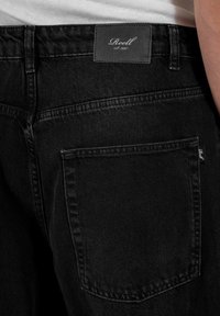 Vaqueros de mezclilla negra con un acabado texturizado, que cuentan con un bolsillo trasero con costura superior y una etiqueta de cuero rectangular en la cintura.