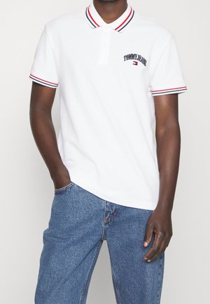 Chemise polo blanche avec col rayé rouge et bleu marine, ornée du logo brodé "TOMMY JEANS" sur la poitrine, accompagnée d'un jean en denim bleu.