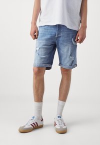 Džínové šortky ve světle modré barvě s opotřebovaným vzhledem, ohrnutým lemem a viditelným třepením. Kombinované s bílými ponožkami a sportovními botami.