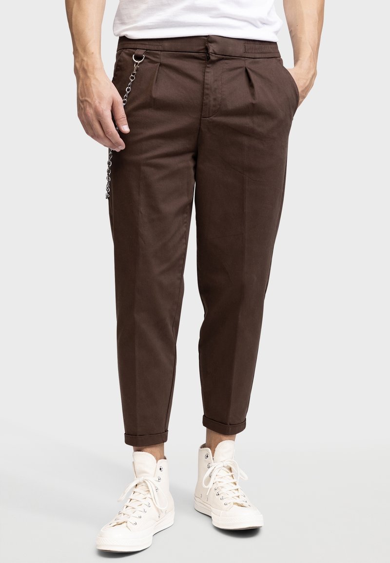 Pantalons chino marron taille haute, avec une devant plissé, des poches latérales et un détail en chaîne accroché à une passant de ceinture, portés avec des baskets montantes blanches.
