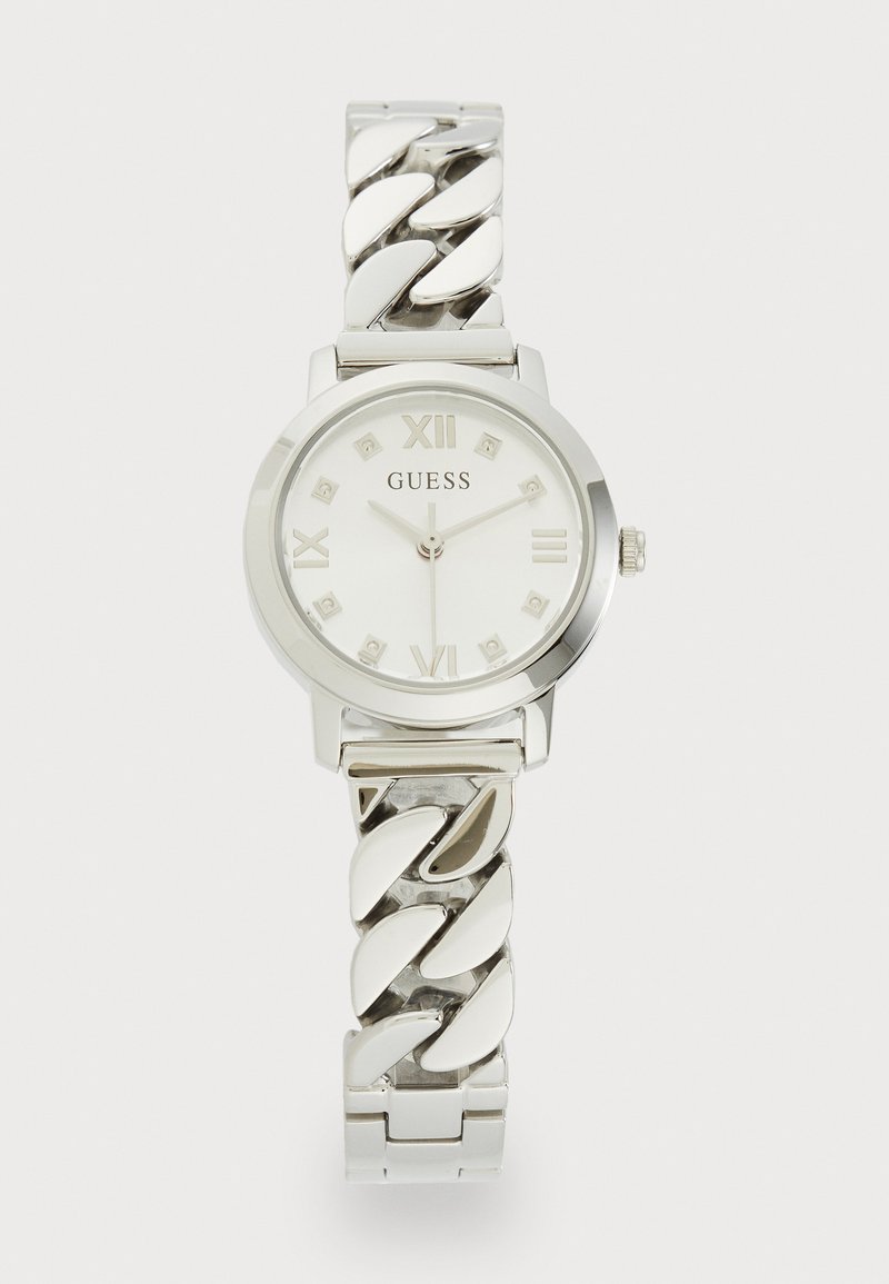 Reloj plateado redondo con una esfera blanca, marcadores de números romanos y una pulsera de cadena enlazada. Presenta pequeñas piedras decorativas en la esfera.