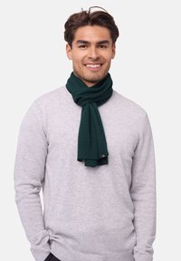 Mørkegrønt strikket tørklæde med ribbet tekstur, vist afslappet omkring halsen på en person iført en lys grå sweater.