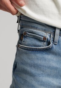 Jeans de mezclilla azul claro con un corte relajado, que presentan remaches en tono cobre, una pequeña etiqueta con el logotipo y un ligero desgaste en la textura de la tela.