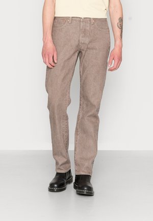 Hombre con vaqueros beige de pierna recta, botas de cuero negras y una camiseta crema, de pie contra un fondo blanco liso.