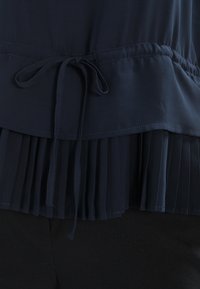 Marinblus i texturerad tyg, med en plisserad nedre fåll och en knytmidja för justerbar passform.