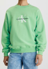 Zelená mikina vyrobená z měkkého materiálu, s bílým potiskem loga "CalvinKleinJeans" na přední straně a žebrovanými manžetami a spodním lemem.