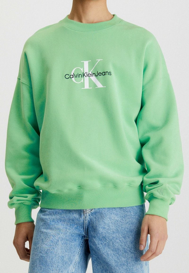 Zelená mikina vyrobená z měkkého materiálu, s bílým potiskem loga "CalvinKleinJeans" na přední straně a žebrovanými manžetami a spodním lemem.