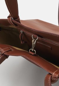 Brown leather handbag with a smooth exterior, contrasting stitching, and a zippered interior pocket. Features metal hardware and a spacious design.