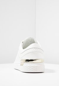 White sneaker with textured upper, gold accent on heel, and rubber sole with diamond pattern. Design features low profile and clean lines.