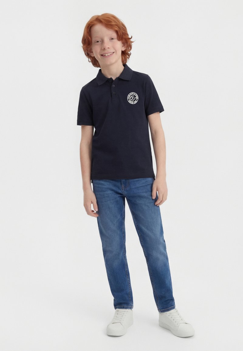 Αγόρι με κοκκινομάλλες που φοράει μπλε πόλο μπλούζα με στρογγυλό λευκό λογότυπο, μπλε τζιν και λευκά αθλητικά παπούτσια, στέκεται και χαμογελά σε λευκό φόντο.