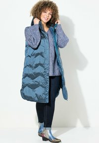 Langer, gepolsterter Weste in Teal mit Chevron-Steppung, über einem strukturierten blauen Pullover getragen. Kombiniert mit dunklen Jeans und metallischen Ankle Boots.