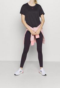 Černé sportovní tričko s malým červeným logem, v kombinaci s tmavými legíny a růžovým svetrem uvázaným v pase. Bílé boty s květinovými akcenty.