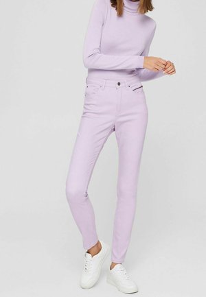 Jeans de tiro alto en color lila con un corte ajustado, cuatro bolsillos y cremallera. Combinados con un suéter de cuello alto en un tono de lila claro y zapatillas blancas.