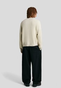 Sweter dziany w kolorze kremowym z prążkowaną fakturą, luźny krój i długie rękawy, w połączeniu z czarnymi spodniami o szerokich nogawkach i ciemnym obuwiem.