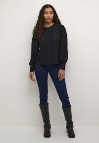 Černý pletený top s texturovaným diamantovým vzorem, balónovými rukávy a kulatým výstřihem, kombinovaný s tmavě modrými skinny džíny a černými kozačkami po kolena.