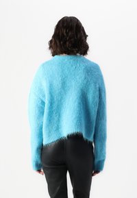 Pull court bleu clair, duveteux, à coupe ample avec des manches longues, porté avec un pantalon noir moulant ; texture douce et détails d'ourlet non fini.