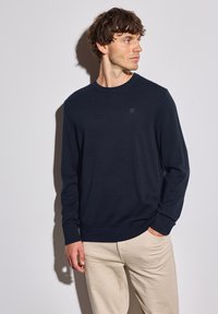 Marineblauer Strickpullover mit Rundhalsausschnitt, gerippten Bündchen und Saum. Kleines Logo-Detail auf der Brust. Kombiniert mit hellbeigen Hosen.