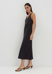 Schwarzes Satin-Midikleid mit V-Ausschnitt, dünnen Trägern und seitlichen Schlitzen. Glatte Textur, lockere Passform und dezentes Muster. Getragen mit braunen Absatzsandalen.