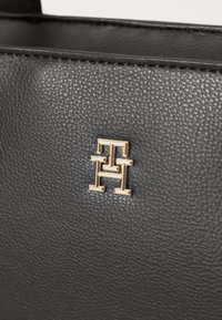 Zwart gestructureerd leer met een gouden logo-detail met de letters 'H' en 'T'. Close-up weergave benadrukt het materiaal en vakmanschap.