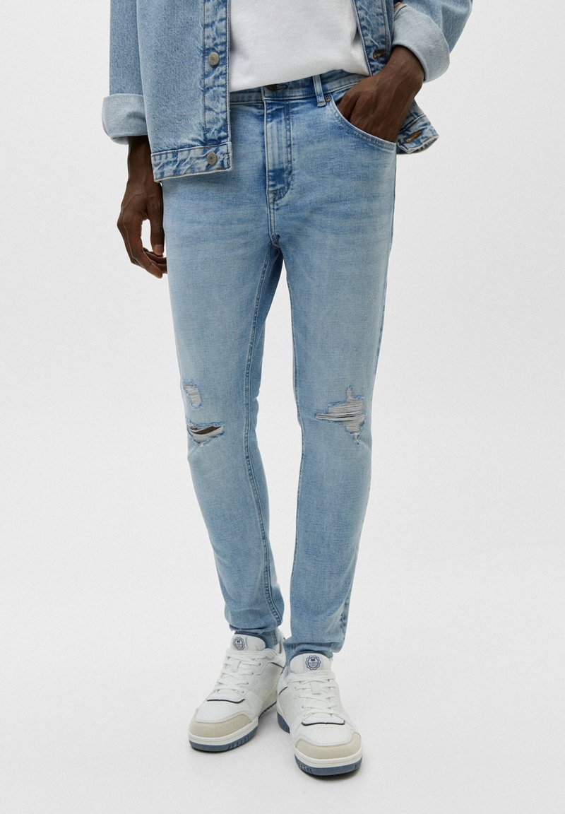 Personne portant un jean bleu clair déchiré, une chemise blanche, une veste en jean et des baskets blanches, une main dans une poche, debout devant un fond uni.