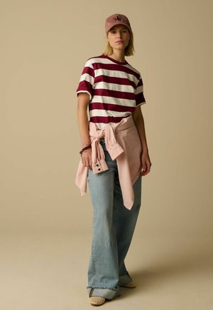 Femme portant un t-shirt rayé bordeaux et blanc, un jean bleu clair, une veste rose nouée à la taille, des chaussures beiges et une casquette rose.