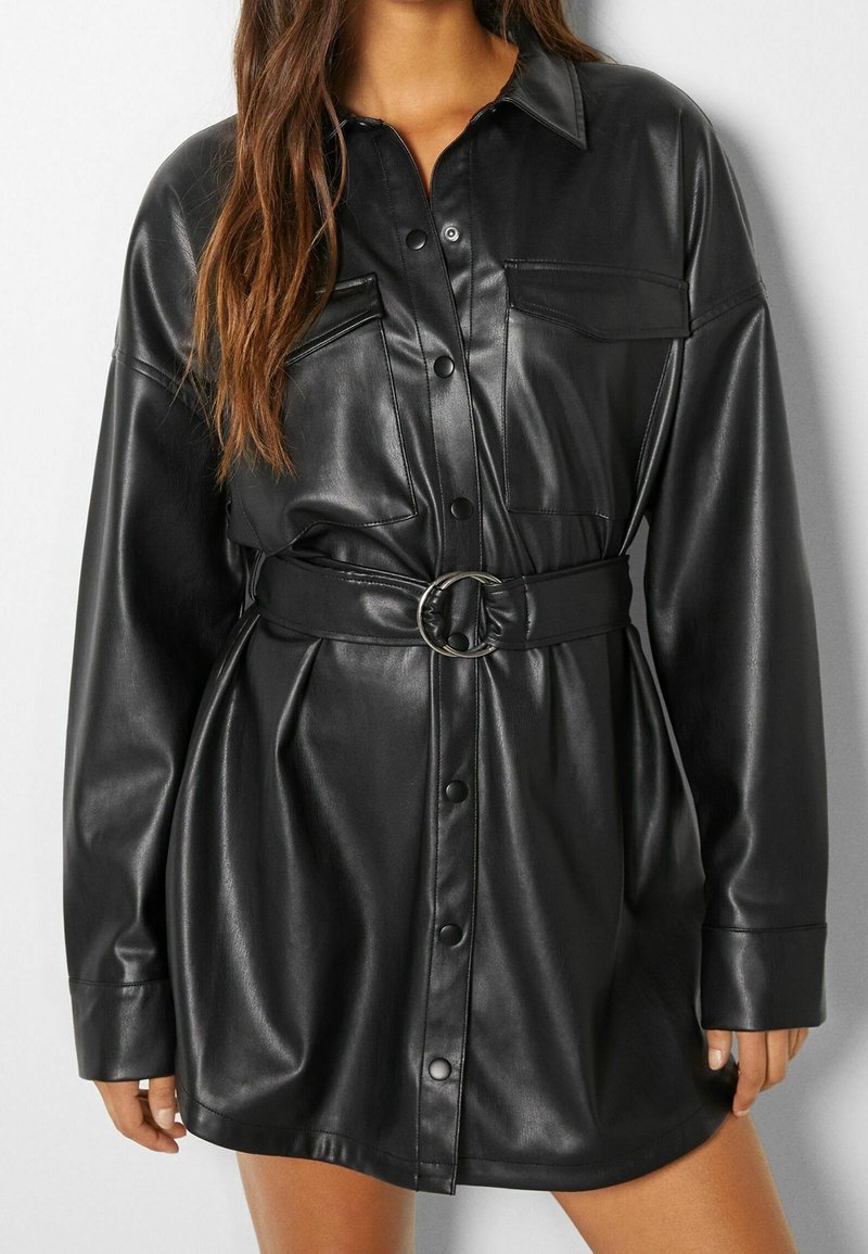Robe chemise en faux cuir noir avec un design à boutons, poches poitrine, manches roulées et une ceinture en tissu avec une boucle circulaire.