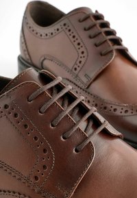 Chaussures de ville en cuir marron avec des détails en brogue, présentant des lacets ronds, une texture lisse et un talon légèrement surélevé.