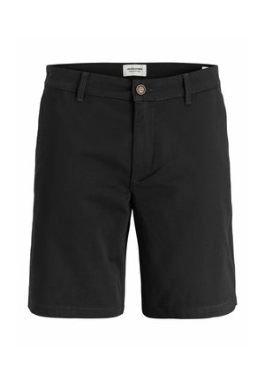 Junge steht mit den Händen in den Taschen, trägt ein weißes kurzärmeliges Polo, schwarze Shorts, weiße Socken und graue Laufschuhe vor einfarbigem Hintergrund.