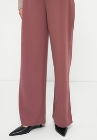 Calças de perna larga numa cor mauve suave, confeccionadas em tecido liso. Apresentam linhas clean, com saltos altos pretos pontiagudos visíveis por baixo.