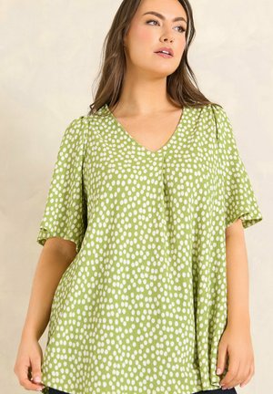 Femme portant une robe verte ample à manches courtes avec des pois blancs, regardant légèrement sur le côté sur un fond uni.