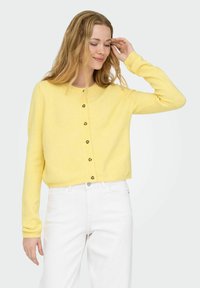 Gul strikket cardigan med rund hals, knappelukking foran og lange ermer. Teksturert stoff vist mot en lys bakgrunn.