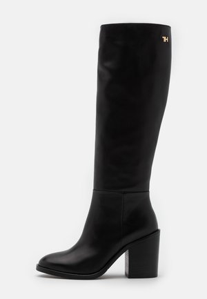 Bottes en cuir noir jusqu'aux genoux avec un talon épais et un bout arrondi. Présente un accent de logo doré près du haut. Texture lisse sur toute la surface.