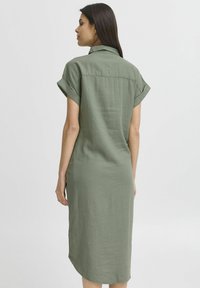Abito in lino verde oliva, slim fit, a maniche corte, con colletto, dettaglio sulla spalla posteriore e orlo leggermente svasato che arriva sotto il ginocchio.